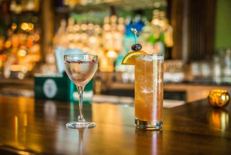 Close up view of two cocktails on a bar with display case of alcohol in the background