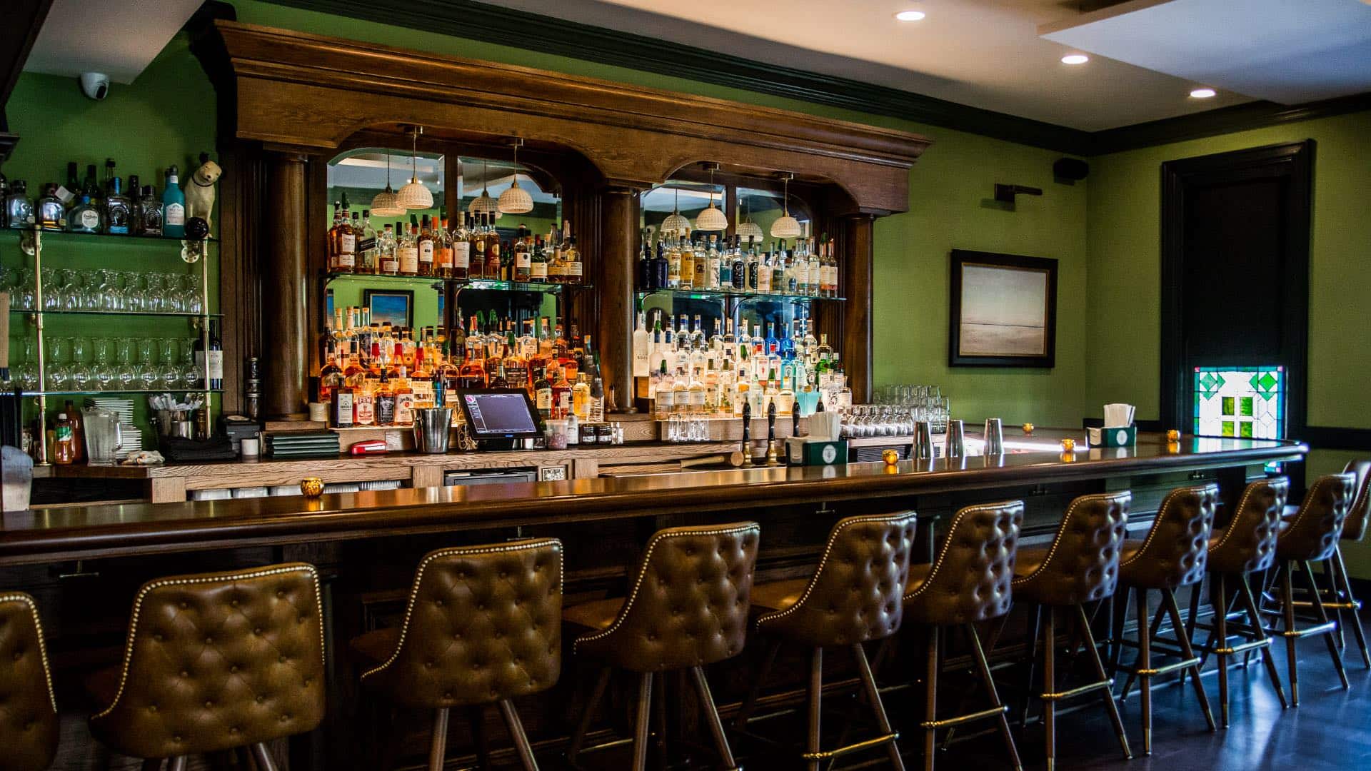 Large wooden bar with leather bar stools and large wooden display case full of bottles of alcohol