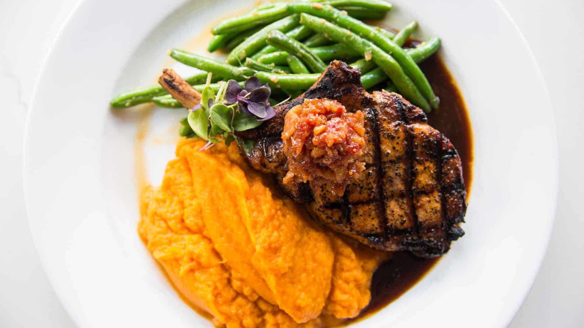 Close up view of grilled pork chop, mashed sweet potatoes, and green beans on white plate