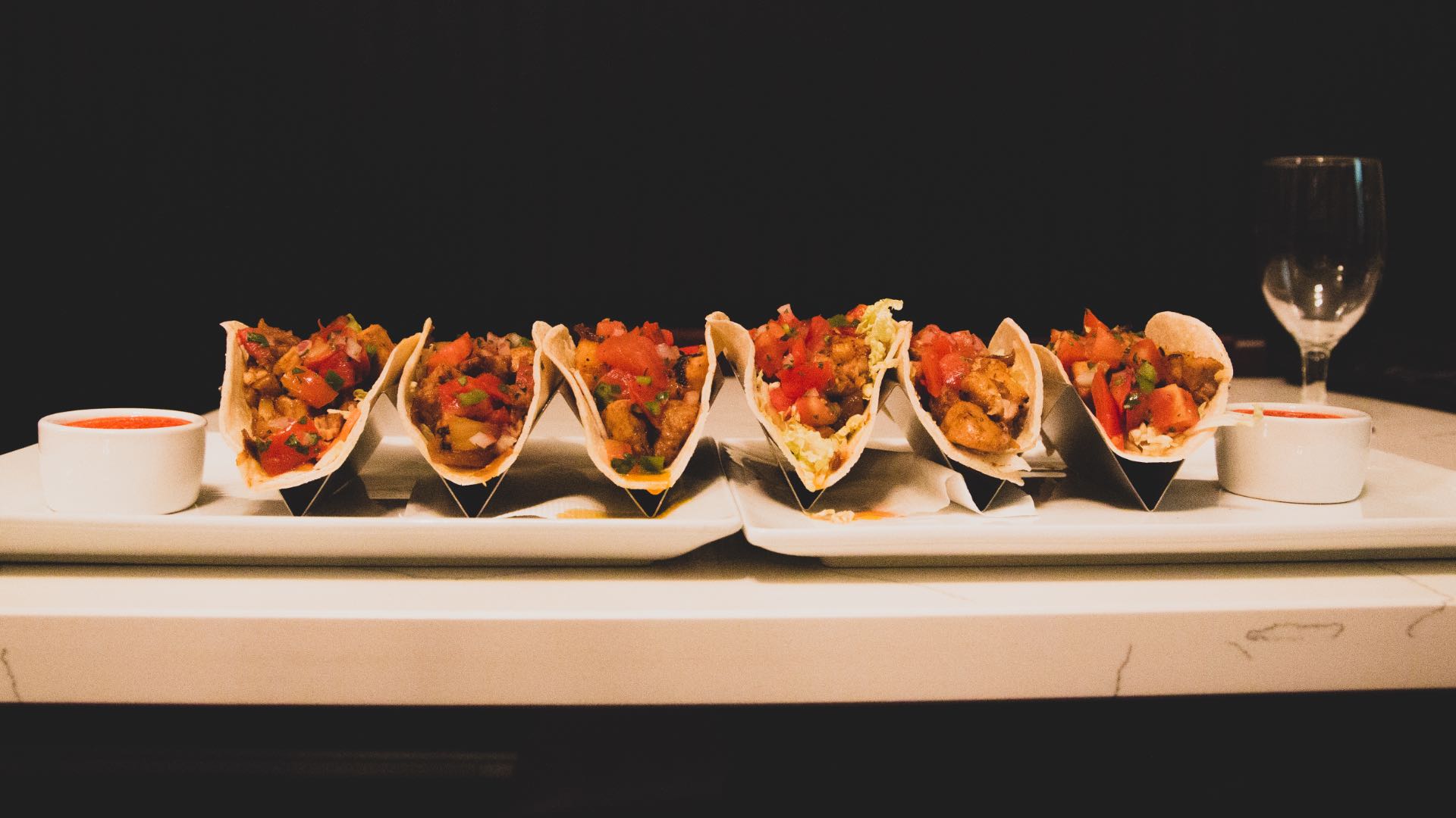 Close up view of two trays of tacos in silver holders on white plates with a dark black background